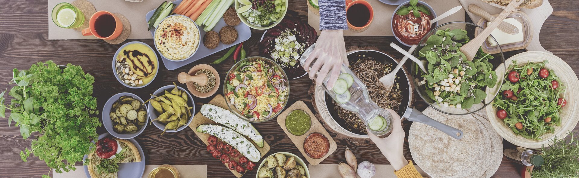 High angle of person pouring water with cucumber to friend's glass above table with vegan food